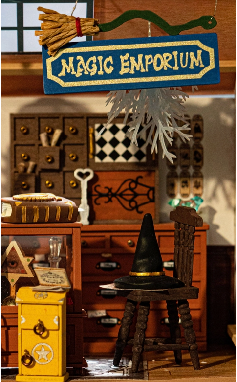 A close up of a witch’s hat sitting on a wooden chair. Behind it is a wall of cubby holes and drawers, holding various magic scrolls and spell ingredients. Above everything is a blue sign reading “magic emporium” in pale yellow text. with a bent broomstick sitting on top of it.