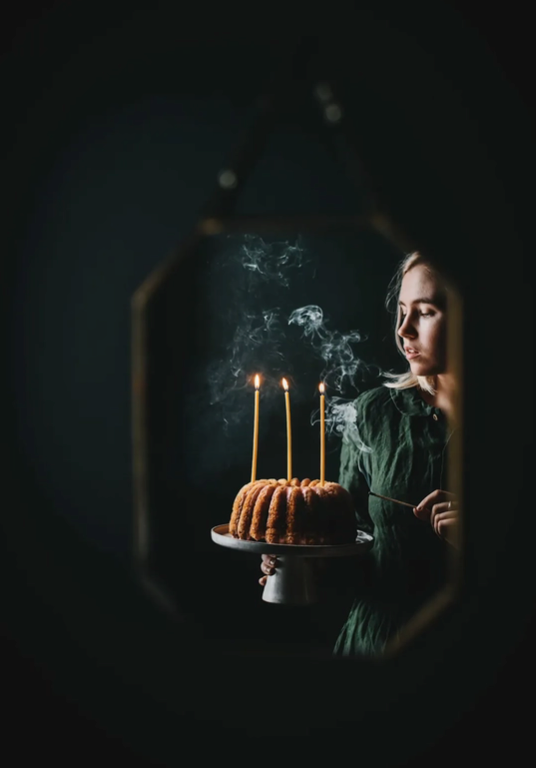 Load image into Gallery viewer, A woman in a green dress holds a cake on a display plate, with lit yellow candles in the cake. Behind the woman is a dark grey background.
