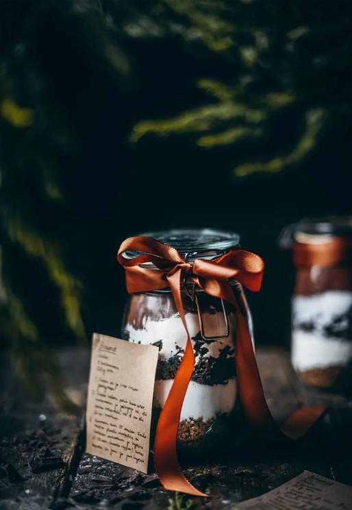A small glass jar with alternating layers of cream and chocolate. A dark orange ribbon bow is tied to the lid. A darkly lit forest is behind the jar.