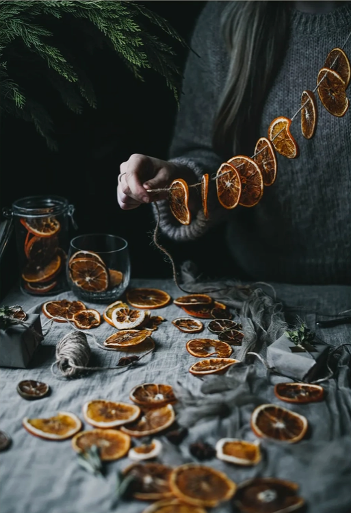 Load image into Gallery viewer, A woman in a grey sweater holds a string with orange slices hanging from it. Below her are more orange slices on a grey cloth, waiting to be added to the string. A darkly lit forest is behind the woman.
