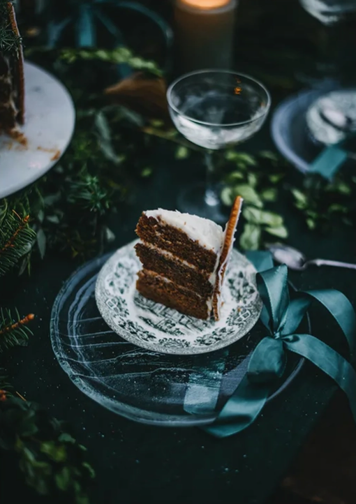 Load image into Gallery viewer, A slice of chocolate cake with white frosting sites on an inricately decorated grey and white plate, with a larger dark grey plate under it. Strewn around the cake are gree sprigs of leaves. In the background are more plates, and wine glasses.
