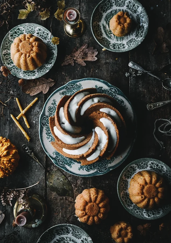 Load image into Gallery viewer, A brown spiral-shaped cake with white frosting sits on a grey and white plate, with a wood table under it. Bext to the cake are smaller bundt-style cakes, on smaller plates. Dried brown leaves are strewn around the table.
