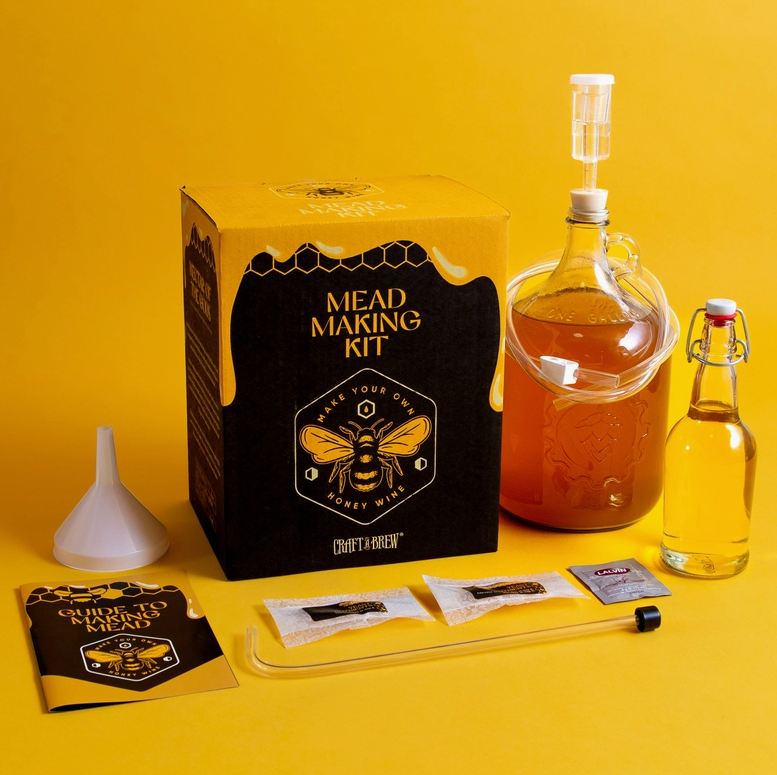 A black and yellow square box on a yellow background. The top of the box depicts honey running down the sides. On the top of the box in yellow text is "mead making kit," with a drawing of a honeybee under it. Next to the box is a one gallon glass jug, filled with a honey-colored liquid, with a stopper in the opening. Beside the jug is a yellow and black booklet, with "guide to making mead" in yellow text. In front of the box are some of the tools from the kit.
