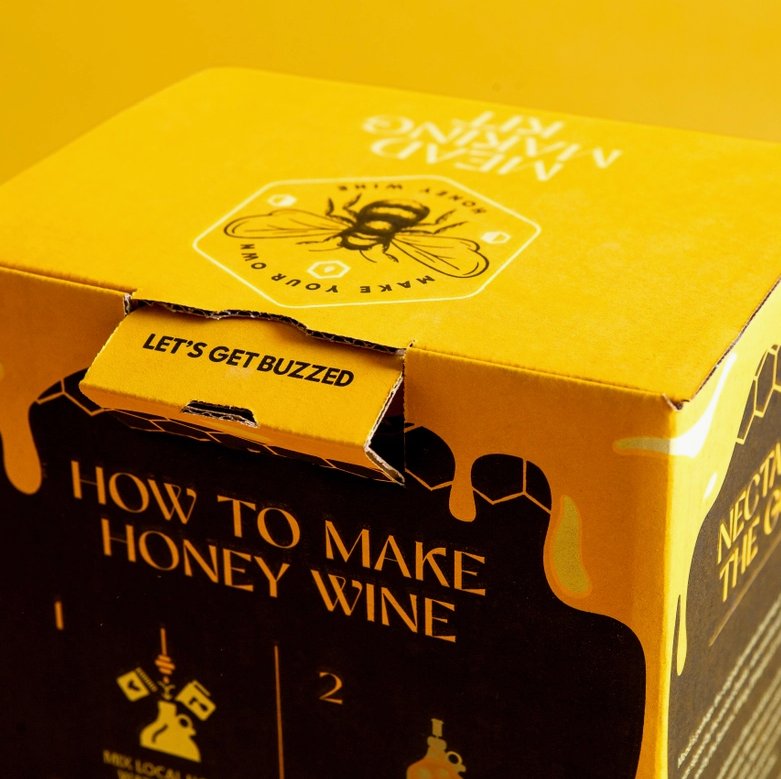 A close up image of a yellow and black box, with a drawing of a honeybee on the top. In black text around the bee is "make your own honey wine."