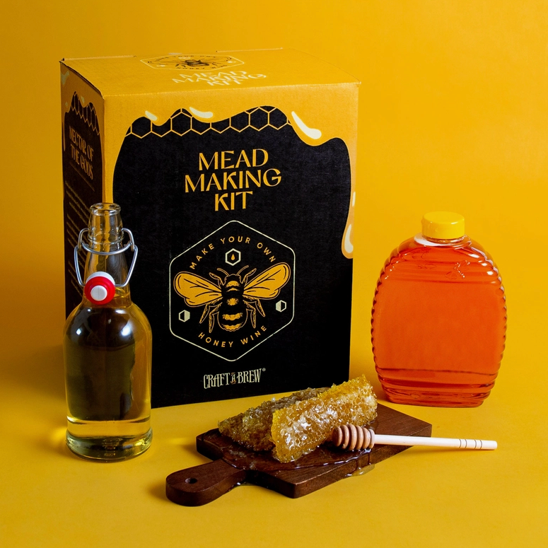 A black and yellow square box on a yellow background. The top of the box depicts honey running down the sides. On the top of the box in yellow text is "mead making kit," with a drawing of a honeybee under it. Next to the box is a plastic jar of honey. In front of the box are a glass bottle filled with clear liquid, and a small wooden cutting board with honeycomb on top of it.