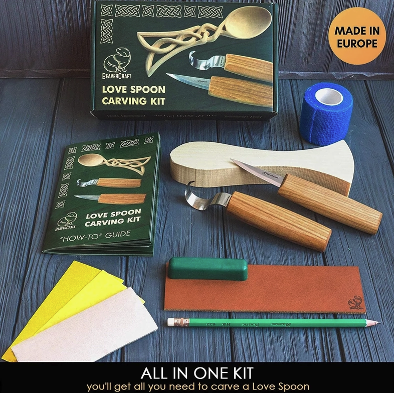 An image of the wood carving kit and its contents on a wooden table. The kit's green box is at the top, with the uncarved spoon and tools under it. Next to the tools is a small book with "how-to guide" printed on the cover. Below the guide are the rest of the tools and pieces for the kit. At the bottom of the image is a black rectangle, with white text saying "All in one kit"