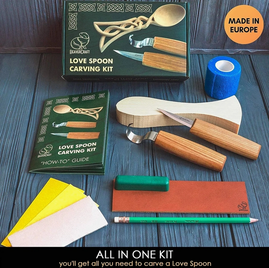 An image of the wood carving kit and its contents on a wooden table. The kit's green box is at the top, with the uncarved spoon and tools under it. Next to the tools is a small book with "how-to guide" printed on the cover. Below the guide are the rest of the tools and pieces for the kit. At the bottom of the image is a black rectangle, with white text saying "All in one kit"