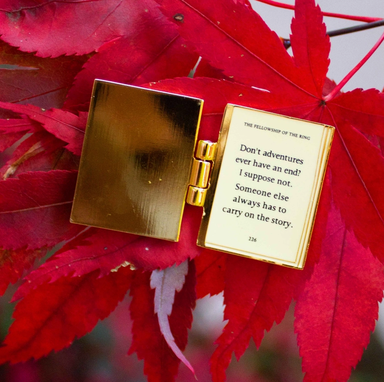 Load image into Gallery viewer, A rectangular enamel pin, opened like a book, on a read leaf. The interior of the pin is gold plated, with a quote from The Lord Of The Rings in black text.
