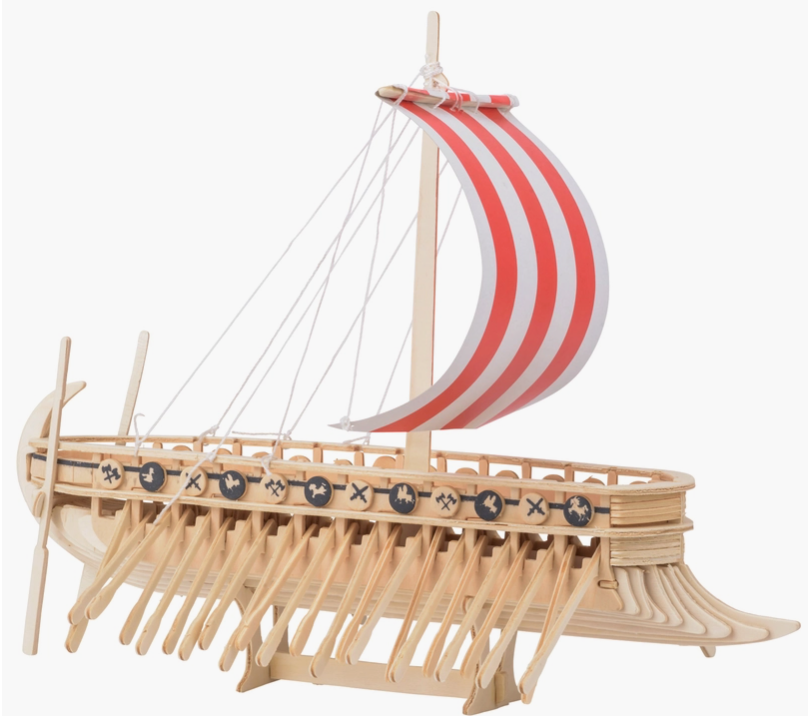 Side view of a miniature wooden Viking ship with a read and white sail, set against a white background. The ship has many oars sticking out the sides, and miniature wooden shields line the edge, with drawings of axes and dragon heads on each.