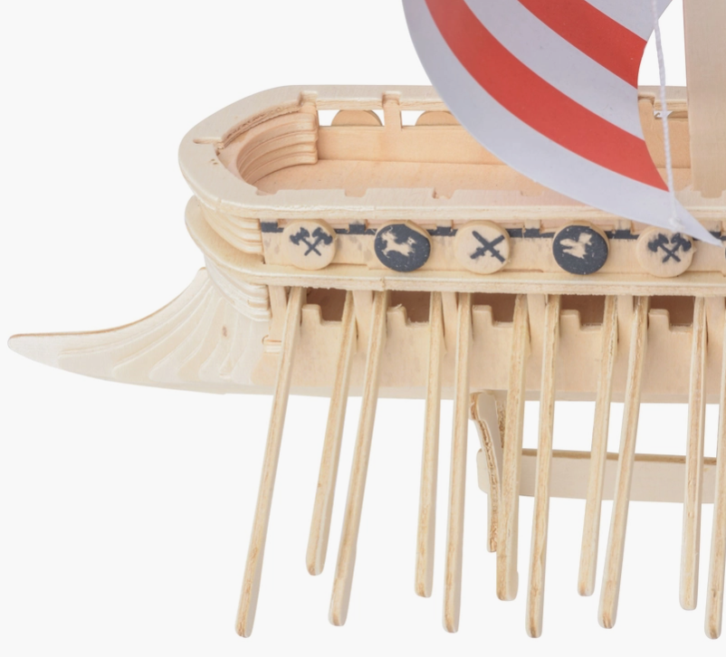 Load image into Gallery viewer, Close up view of the aft section of a miniature wooden Viking ship with a read and white sail, set against a white background. The ship has many oars sticking out the sides, and miniature wooden shields line the edge, with drawings of axes and dragon heads on each.

