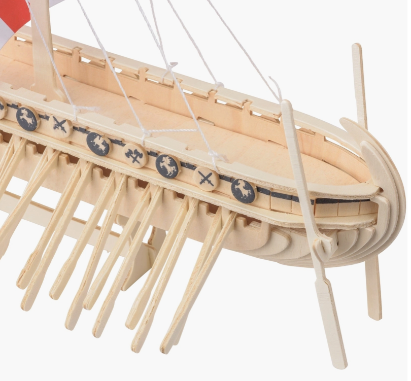 Close up view of a miniature wooden Viking ship, set against a white background. The ship has many oars sticking out the sides, and miniature wooden shields line the edge, with drawings of axes and dragon heads on each.