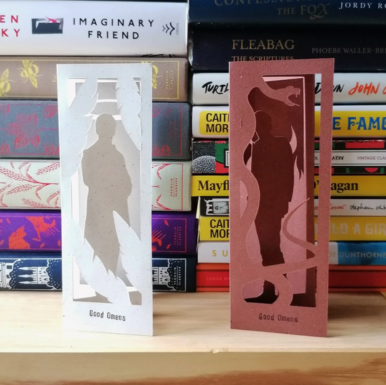 An image of two bookmarks, one red and one white, standing in front of a pile of books. The red bookmark is cut to depict Crowley, and the white is cut to depict Aziraphale, the two main characters from the series "Good Omens."