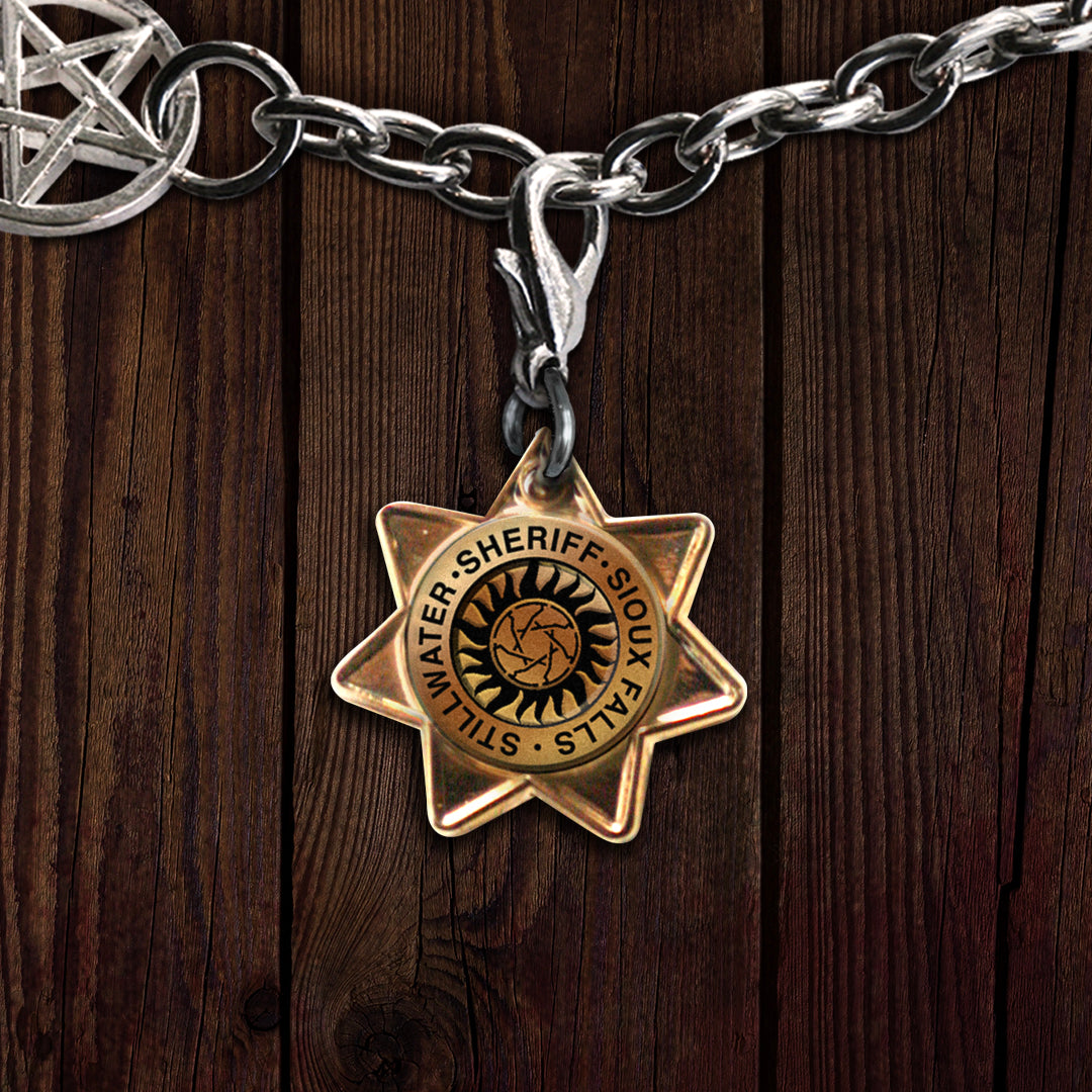 A brass charm in the shape of a Sheriff's badge. On the badge is a small anti-possession symbol encircled by the words SHERIFF - SIOUX FALLS - STILLWATER. The charm is on a silver bracelet/keychain clip. 