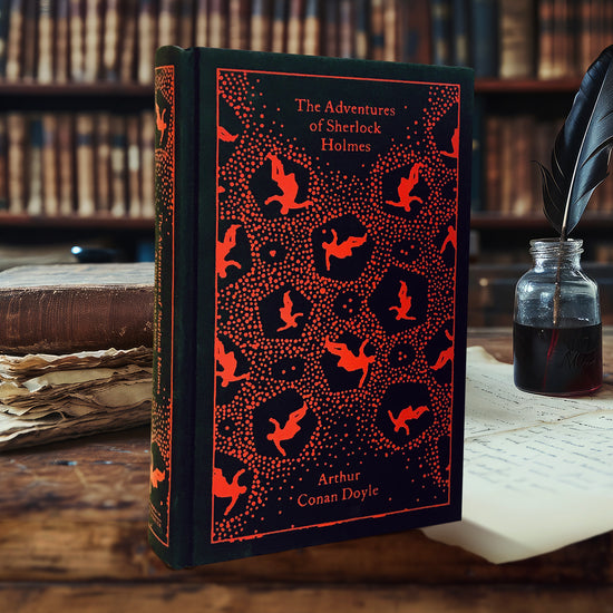 A black hardcover book on a wooden desk. On the front cover are silhouettes of a person falling, in a repeating pattern. Red text at the top says "The adventures of Sherlock Holmes." In the background are shelves lined with reference books.