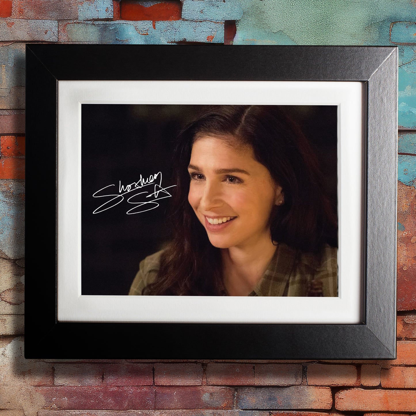 A black framed photograph against a colorful brick wall. The photograph depicts actor Shoshannah Stern, dressed as the character Eileen Leahy from the series "Supernatural." Shoshannah is wearing a green jacket, with a bright smile on her face. In the top left corner is Shoshannah's autograph in white ink.