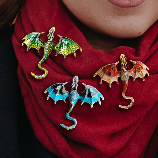Close up view of a model wearing a red scarf. Attached to the scarf are three brooches in the shapes of dragons. One dragon is red, one is blue, and one is green.