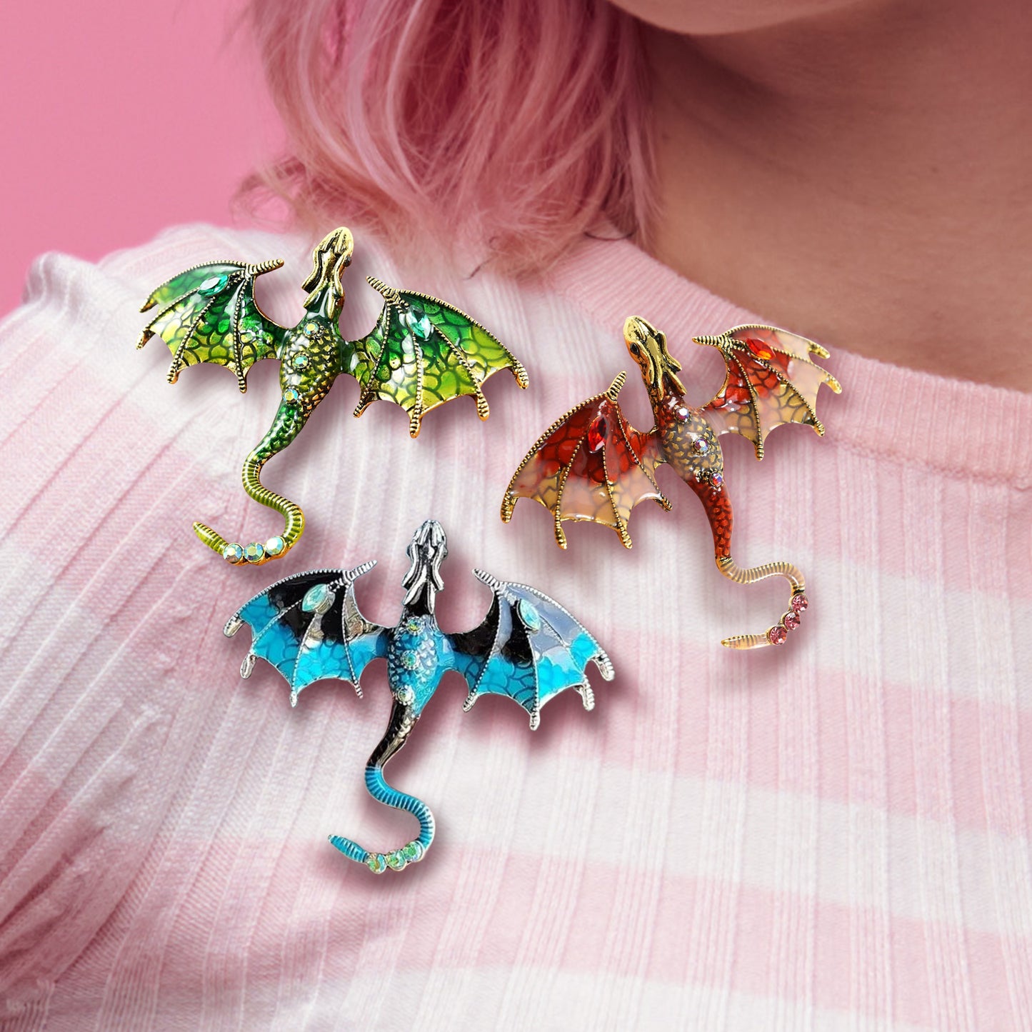 Close up of a model's shoulder, in a pink and white striped shirt. Attached to her shirt are three brooches in the shapes of dragons in flight. One dragon is green, one is red, and one is blue.