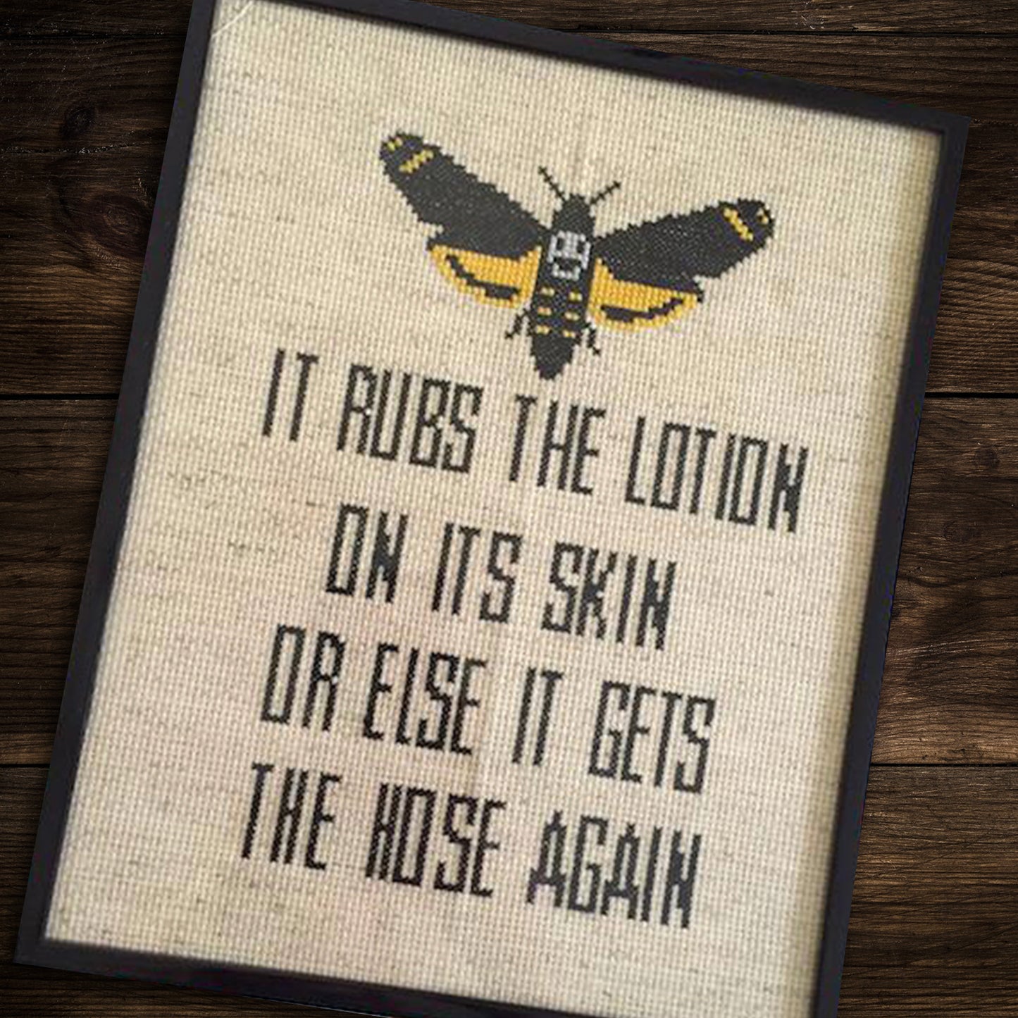 A beige cross-stitch kit, in a black frame, against a dark wooden table. At the top is a yellow and black moth, with a white skull image in its center. Beneath the moth is black text saying "It rubs the lotion on its skin or else it gets the hose again."