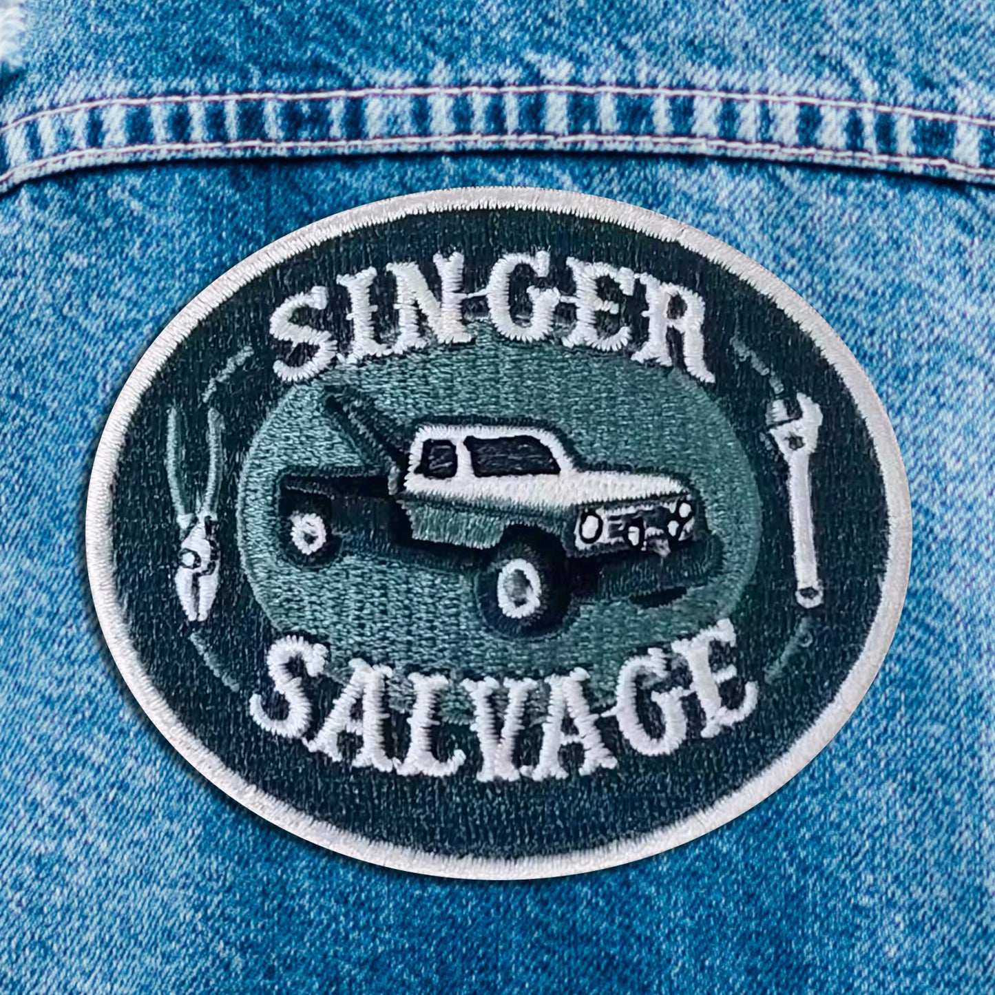Close up of a blue and white iron on patch. The patch depicts a tow truck in the center, with a wrench and pliers on each side. White text says "singer salvage."