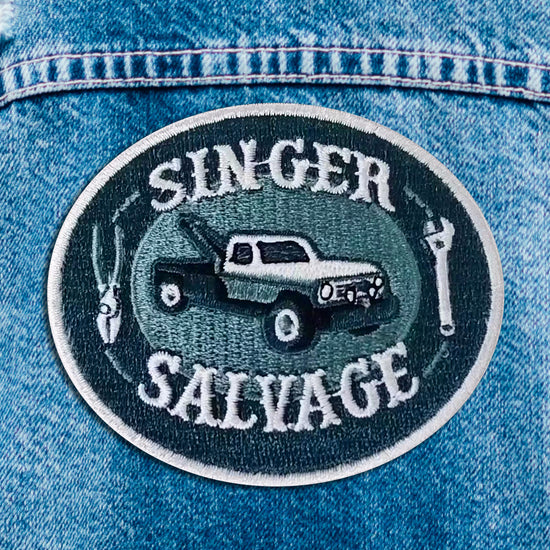 Close up of a blue and white iron on patch. The patch depicts a tow truck in the center, with a wrench and pliers on each side. White text says "singer salvage."