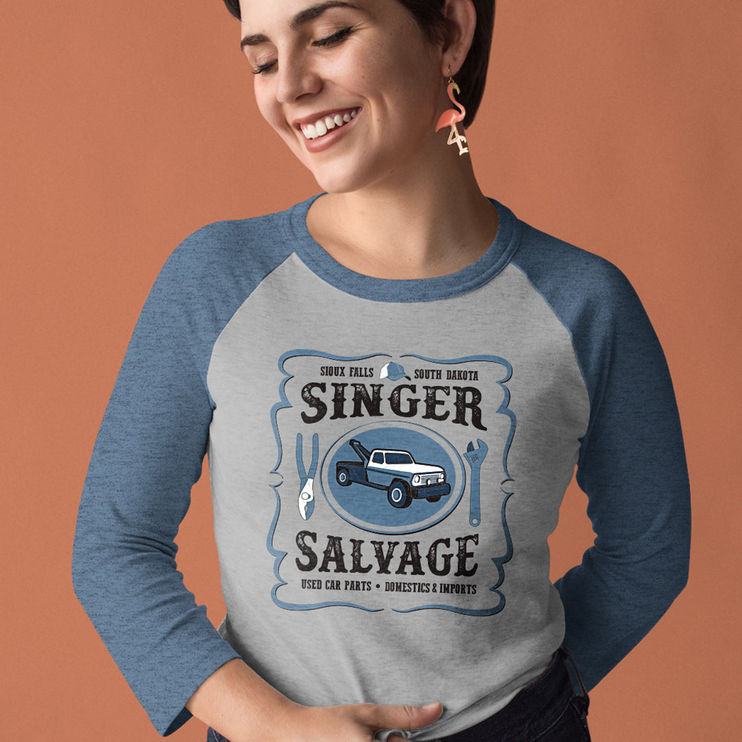 A female model wearing a gray raglan t-shirt with slate blue long sleeves, against a terra cotta colored background. The design on the front of the shirt says "Sioux Falls * South Dakota - SINGER SALVAGE - Used Car Parts * Domestics & Imports" with a tow truck in the center and two tools on either side.