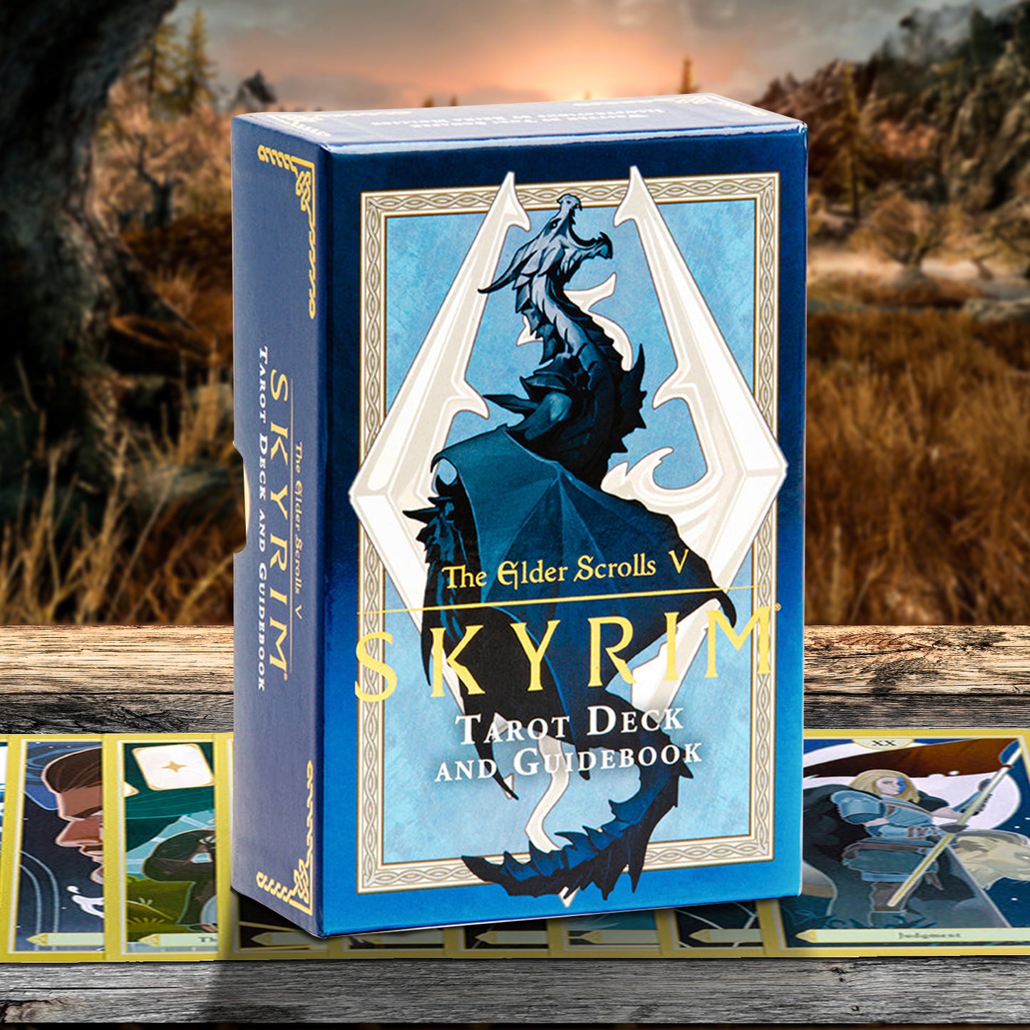 The Elder Scrolls V: Skyrim Tarot Deck and Guidebook box in blue with gold and white artwork, displayed on a wooden surface with cards spread out in front against a blurred outdoor fantasy landscape background.