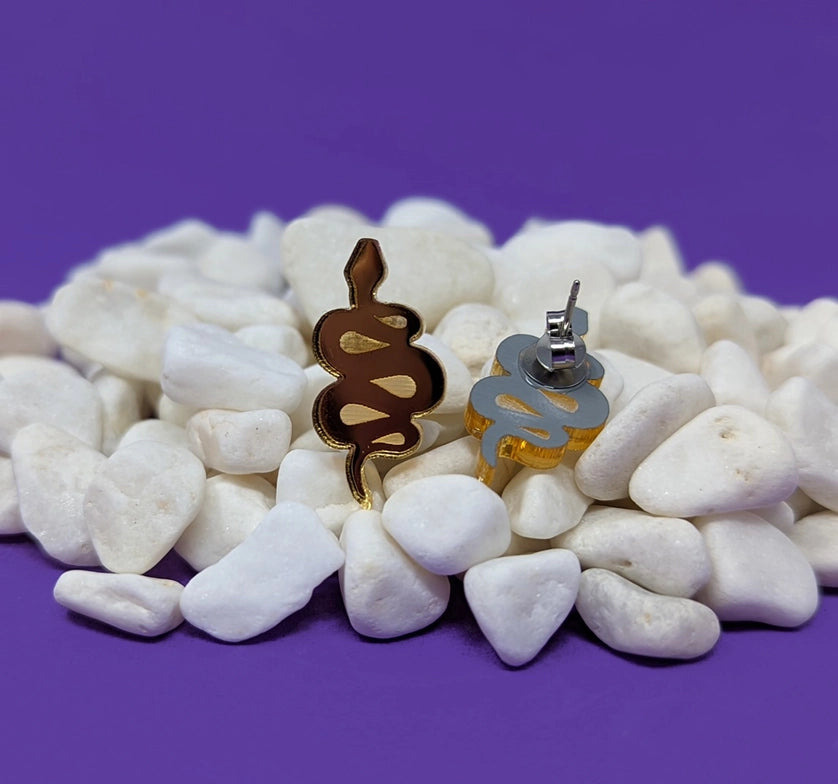 A pile of small white stones against a purple background. In the middle of the pile are two small gold earrings, in the shape of coiled snakes.