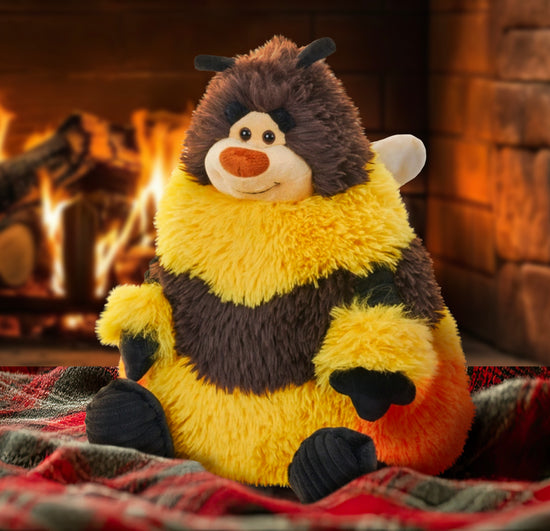 A bee plushie sitting on a red and green plaid blanket. The bee has yellow and brown striped, with white wings on his back. At his bottom is a patch of orange, to represent pollen. The bee has black hands and feet, and a smile on his face. Behind the bee is a roaring fireplace.