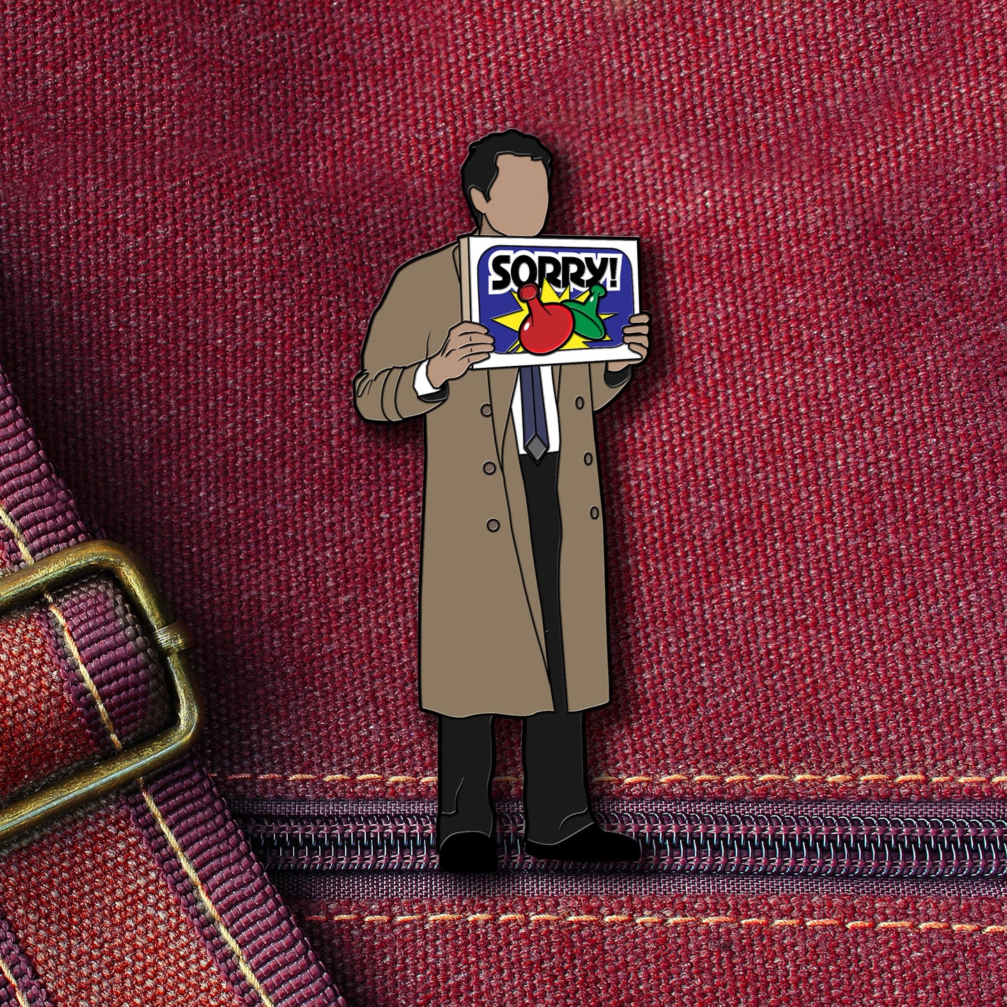 An enamel pin depicting Castiel from the TV series "Supernatural." Castiel is wearing his trademark tan overcoat, and carrying a "Sorry!" board game box. The pin is attached to a maroon canvas bag.