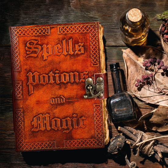 A reddish-brown, leathery journal on a wooden table. The front cover has raised text saying "spells, potions, and magic." At the right side is a metal locking clasp, securing the book shut. Ragged, antique-looking pages are visible at the edge. Next to the journal are small glass vials, bundled twigs, leaves, and dark berries.