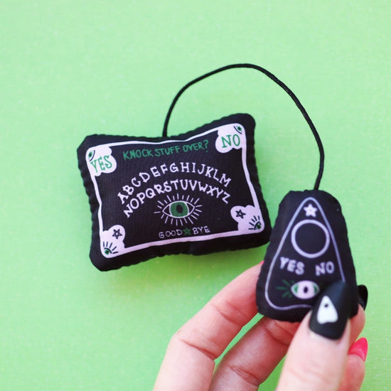 A small black cat toy on a green background, held in a model's fingers. The toy is shaped like a tiny Ouija board with an attached planchette.