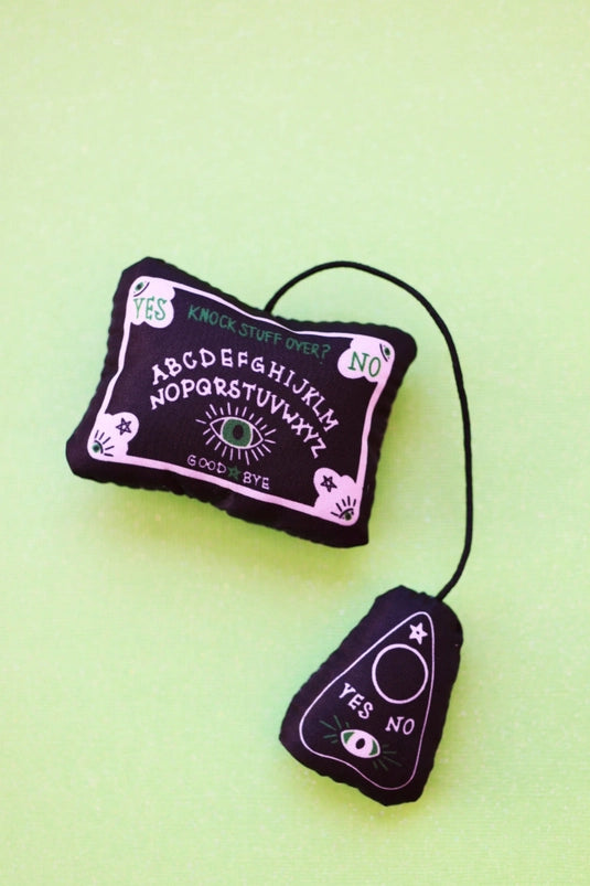 A small black cat toy on a green background. The toy is shaped like a tiny Ouija board with an attached planchette.