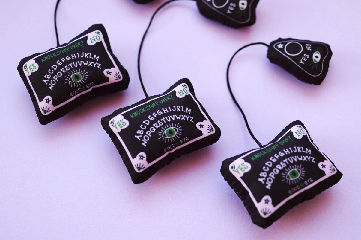 Small black cat toys on a purple background. The toys are shaped like tiny Ouija board with attached planchettes.