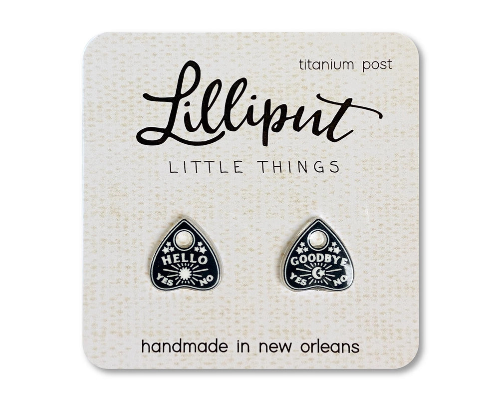 A pair of black earrings depicting tiny planchettes. The left earring has text saying "hello, yes, no" with a sun and stars. The right earring has text saying "goodby, yes, no" with a crescent moon and stars. The earrings are inserted into a white cardboard backing, with black text saying "lilliput, little things."