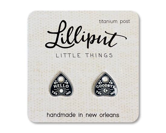 A pair of black earrings depicting tiny planchettes. The left earring has text saying "hello, yes, no" with a sun and stars. The right earring has text saying "goodby, yes, no" with a crescent moon and stars. The earrings are inserted into a white cardboard backing, with black text saying "lilliput, little things."