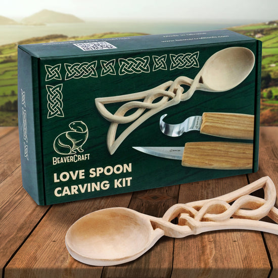An image of a green box on a wood table, in front of farmland, with a carved wooden spoon on the box top. The shape of the handle is similar to a Celtic knot pattern. Under the spoon are two small wood carving tools. On the top of the box are line drawings in a repeating Celtic knot pattern. At the bottom is white text saying "Love spoon carving kit." In front of the box is the carved spoon.