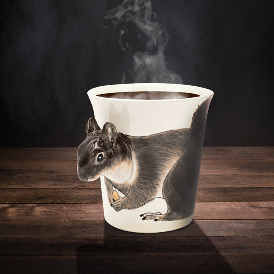 A ceramic mug on a wooden table. The mug depicts a squirrel drawn on the front, with the squirrel's head sticking out as the mug handle. The mug is filled with a brown liquid, and steam is rising from the top.
