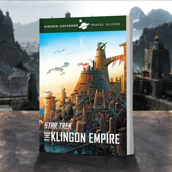 A travel guide book on a stone ledge. The cover depicts a Klingon city from the Star Trek universe. Tan stone buildings with red banners are clustered in the middle. Several Klingon warships are in the sky. At the top is a green banner with white text saying "hidden Universe Travel Guides," and a drawing of the planet Saturn sits in the center. At the bottom is white text saying "Star Trek: The Klingon Empire." Behind the book is a Klingon city.