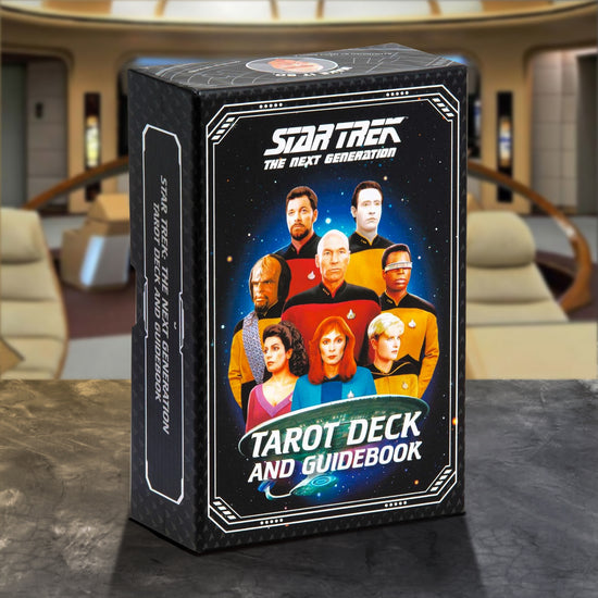 A black cardboard box on a marbled table. The front of the box features drawings of characters from Star Trek: The Next Generation, with a starry background behind them and the USS Enterprise under them. White text across the top and bottom says "Star Trek The Next Generation Tarot Deck and Guidebook." Behind the box is the bridge of the USS Enterprise 1701-D.