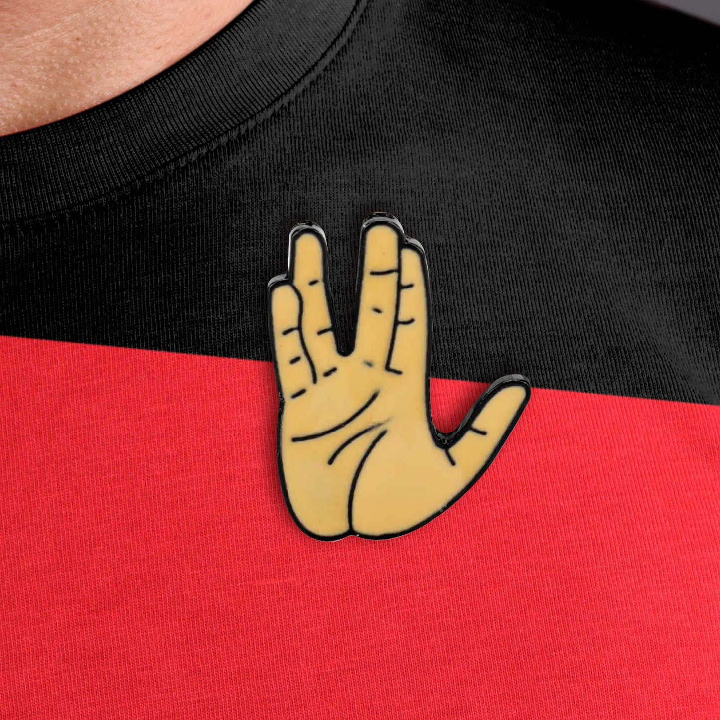 Close up of an enamel pin in the shape of the Vulcan Salute, attached to a red and black shirt. The Salute depicts an upright hand, with the fingers split into a V shape.