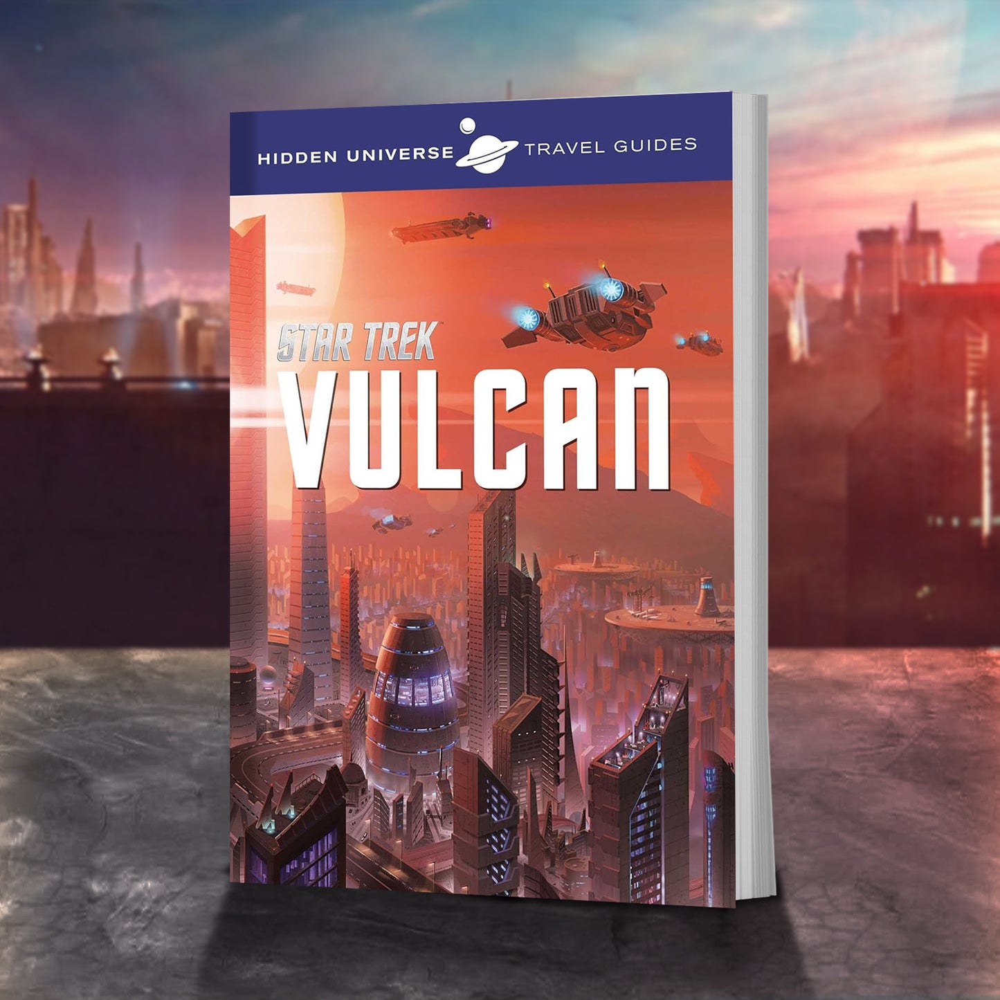 A travel guide book on a stone ledge. The cover depicts a Vulcan city from the Star Trek universe. Futuristic buildings reach into the red sky, with spaceships flying between them. At the top is a purple banner with white text saying "hidden Universe Travel Guides," and a drawing of the planet Saturn sits in the center. In the center is white text saying "Star Trek: Vulcan." Behind the book is a Vulcan city.