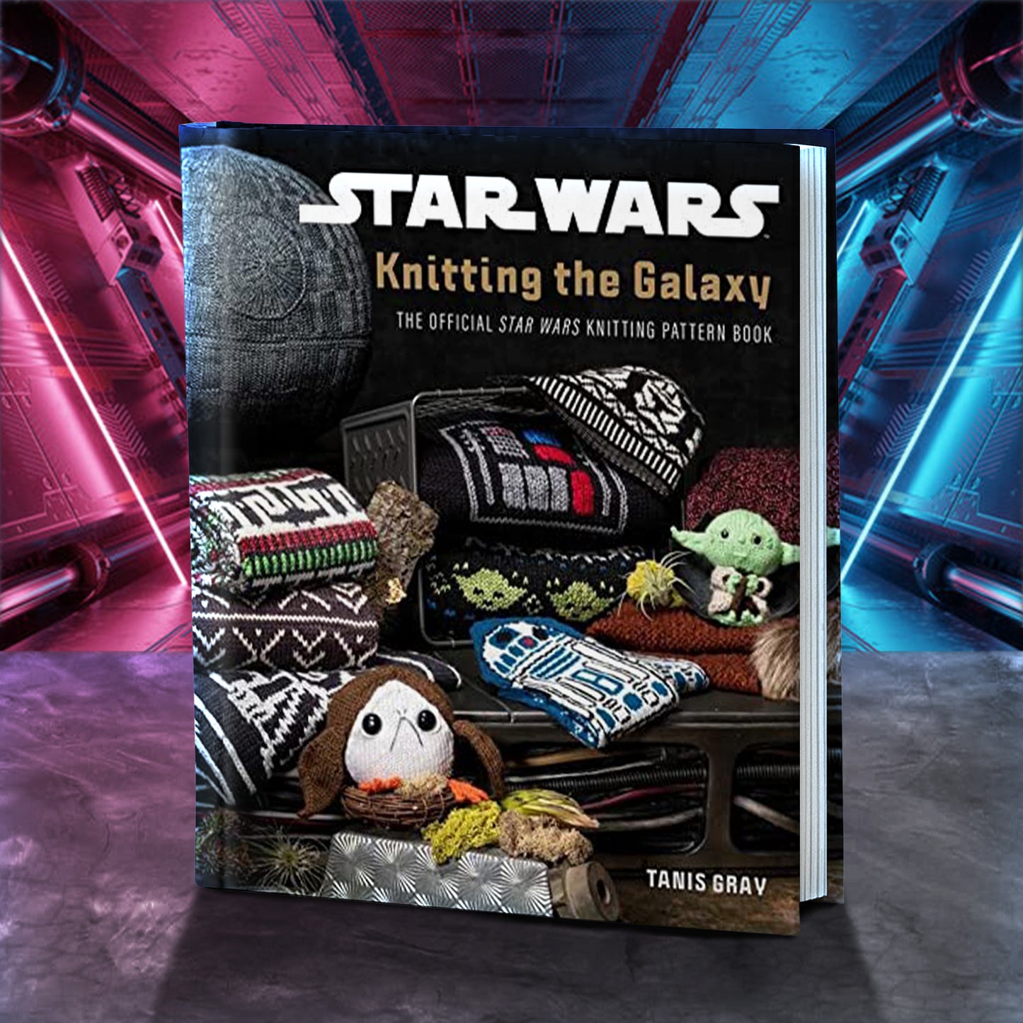 A book standing on a marble table. On the cover are various characters and ships from the Star Wars movies in the forms of knit scarves and sweaters. The book title “Star Wars: Knitting the Galaxy” is across the top in white and gold text. Behind the book is a spaceship corridor, lit in red and blue.