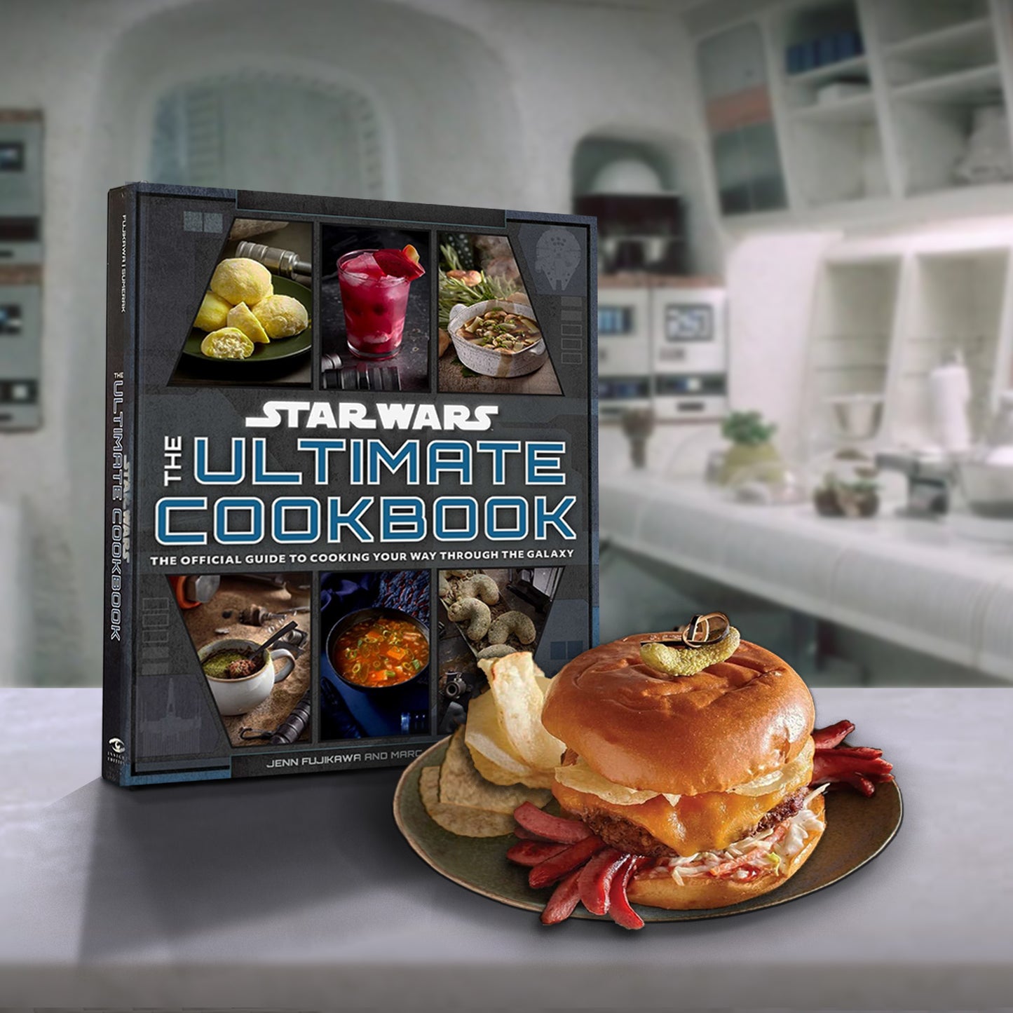 An gray book white marble table. On the front in white and blue text is "Star wars, the ultimate cookbook. The official guide to cooking your way through the galaxy." At the top and bottom are images of some of the recipes in the book. Next to the book is a cheeseburger on a plate. Behind the book is a futuristic-looking kitchen with white surfaces and appliances.