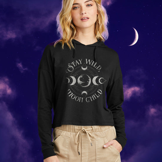 A female model wearing a black long sleeve T-shirt with silver text saying "Stay Wild Moon Child." There is a depiction of the Moon in the center, with crescent Moon phases at top, bottom, left, and right of the center. There are thin circles around the moon phases, connecting them to each other. Behind the model is a nighttime sky.