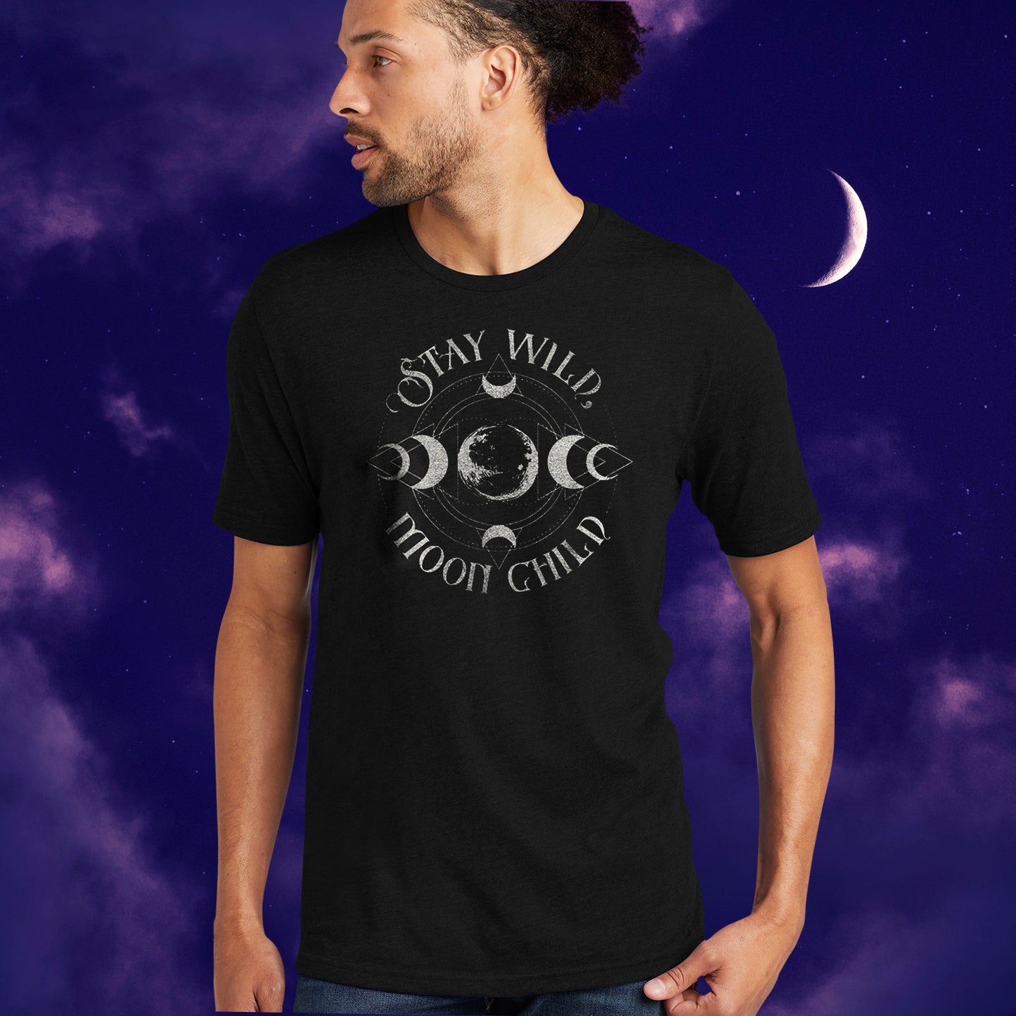 A male model wearing a black T-shirt with silver text saying "Stay Wild Moon Child." There is a depiction of the Moon in the center, with crescent Moon phases at top, bottom, left, and right of the center. There are thin circles around the moon phases, connecting them to each other. Behind the model is a nighttime sky.