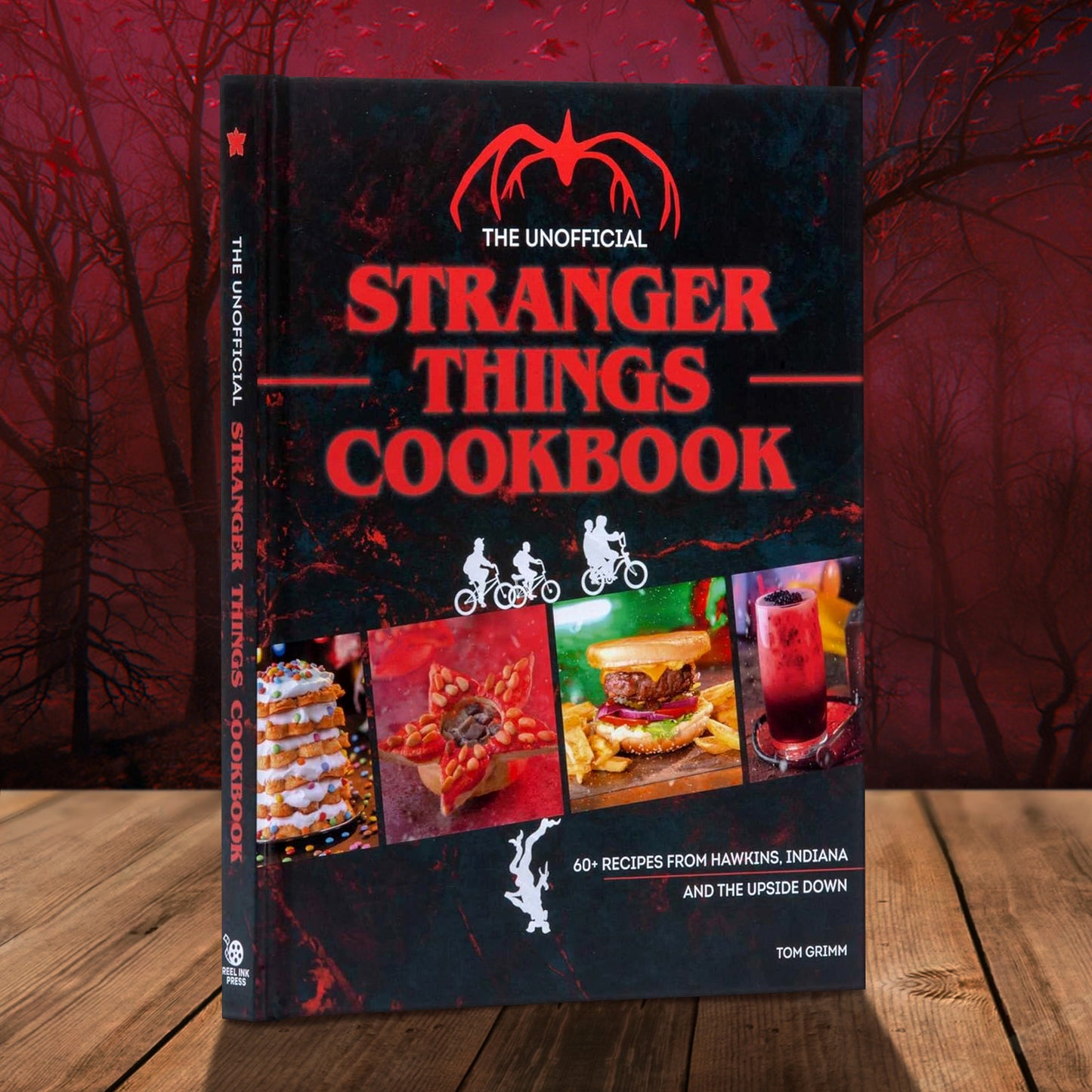 A red and black book on a wood table. Red and white text at the top says "The unofficial stranger things cookbook." A red silhouette of a menacing creature sits above the title. Images of recipes from the book stretch across the bottom, with white silhouettes of kids on bicycles riding across. Below them is an upside down white silhouette of a demogorgon from the series. Behind the book is a dark forest in red and black tones.