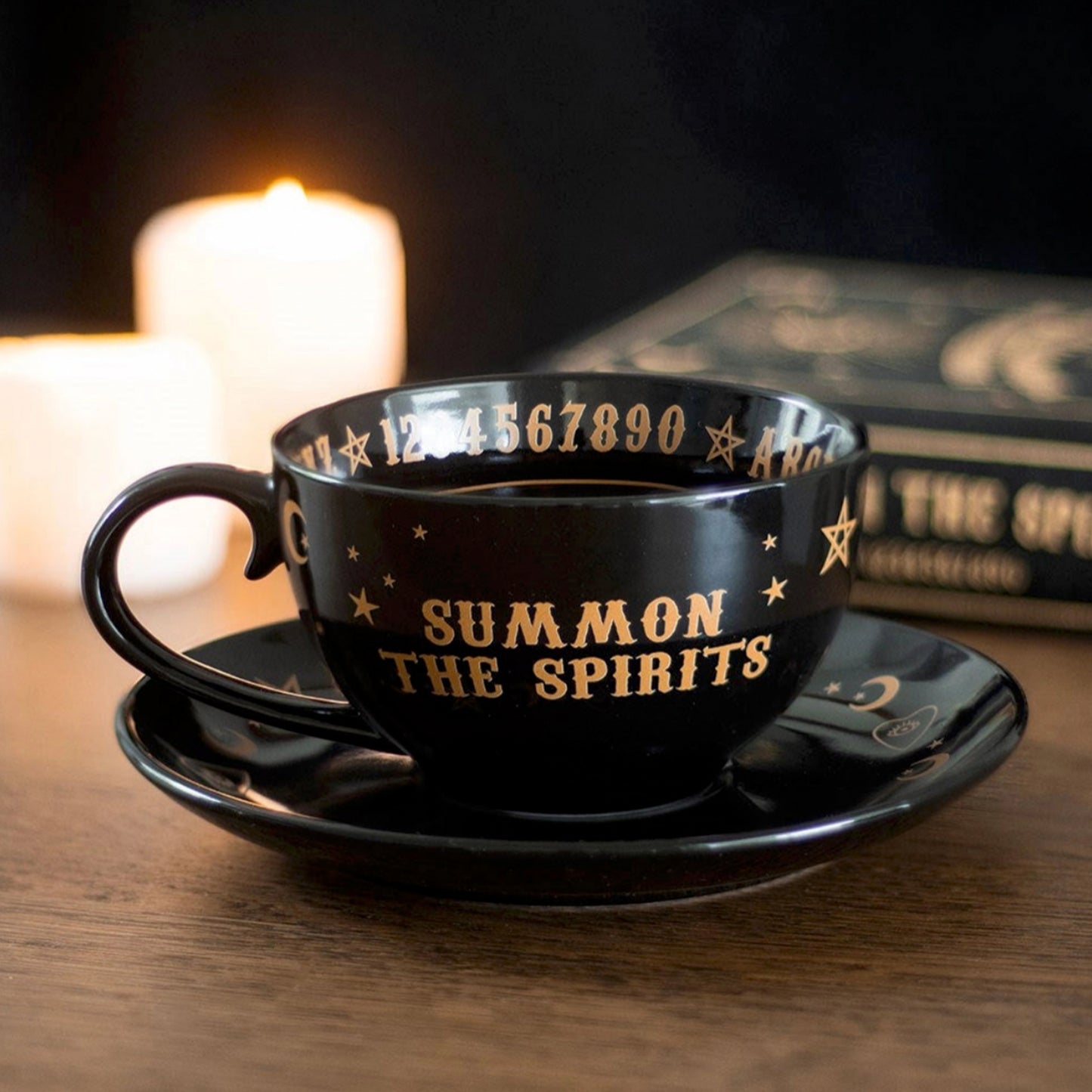 Black teacup and saucer with 'Summon the Spirits' text on a wooden surface with candles and books in the background.
