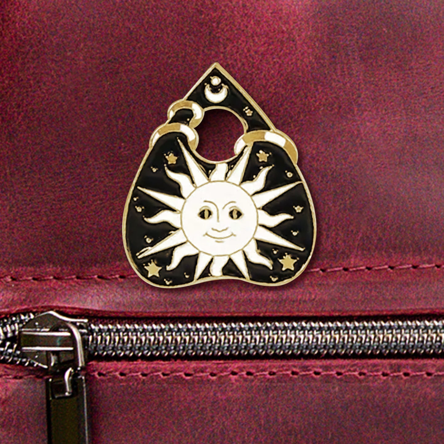 A close-up photo of a black-and-gold enamel pin in the shape of a planchette. The pin depicts a large white sun in a tarot-like art style, surrounded by small gold stars.