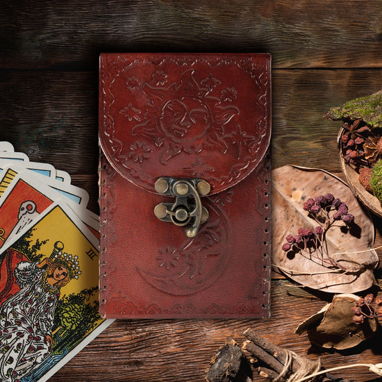 A reddish-brown leather tarot card holder with a brass closure clasp. The leather is embossed with a tarot-art-style sun on the front flap, and a moon on the bottom front.