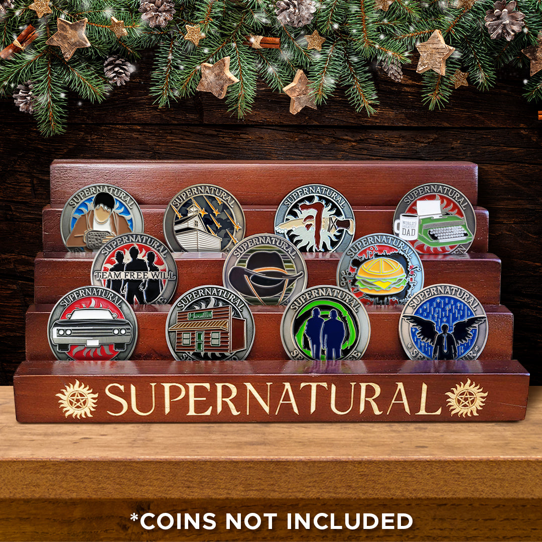 A wooden coin display rack with five tiers, on a table. Behind the rack is the bottom of a Christmas wreath. Sitting on the rack tiers are 11 different Supernatural Challenge Coins. Yellow text on the bottom tier says "supernatural" with the anti-possession symbol at each side. At the bottom of the image is white text saying "coins not included."